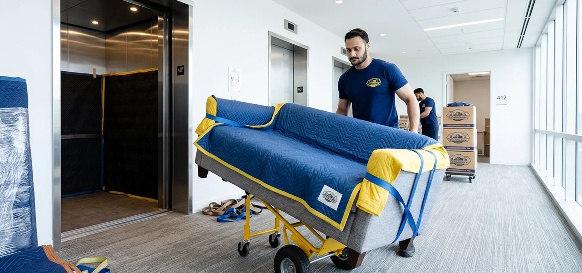 Economical movers transporting a protected sofa through an apartment building hallway near an elevator